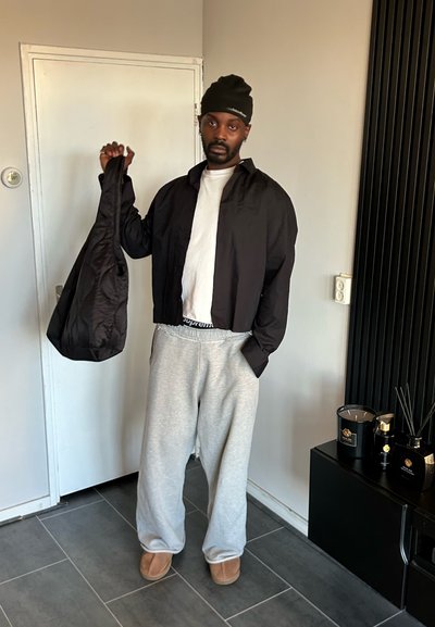 Chaqueta negra corta, camiseta blanca, pantalones deportivos grises y zapatillas de estar por casa color beige. La persona sostiene una bolsa negra. Ambiente interior minimalista con velas.