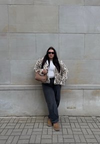 Pelliccia sintetica a pois, top bianco, jeans neri a gamba larga, stivali alla caviglia marroni e borsa a tracolla marrone intrecciata. Sfondo grigio.