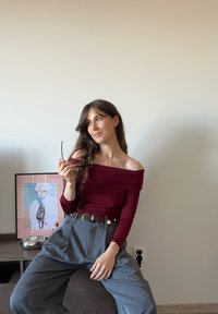 Top bordeaux con spalle scoperte e tessuto arricciato, abbinato a pantaloni grigi a vita alta. Superficie in legno con un'opera d'arte e un oggetto circolare.