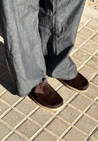 Rjavi copati obloženi s krznom in mehko teksturo, z šivanim vzorcem in rjavimi vezalkami, kombinirani z sivimi, širokimi hlačami na tlakovcu.