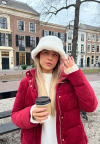 Manteau matelassé rouge, chapeau seau blanc et duveteux, pull léger. Tient une tasse à café à couvercle noir avec un manchon en papier. Cadre extérieur avec des bâtiments.