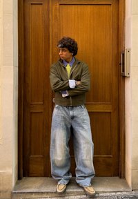 Ung mand med krøllet hår står med armene krydset foran en trædør, iført en grøn jakke, et gult slips og posede jeans.
