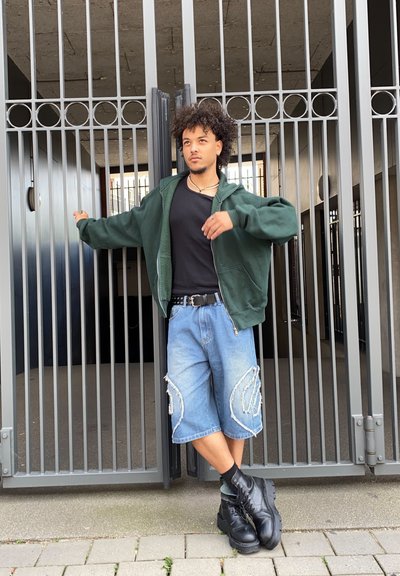 Chaqueta con cremallera verde, camiseta negra, pantalones cortos de mezclilla azules de gran tamaño con costura blanca y botas negras de cordones. De pie frente a una puerta.