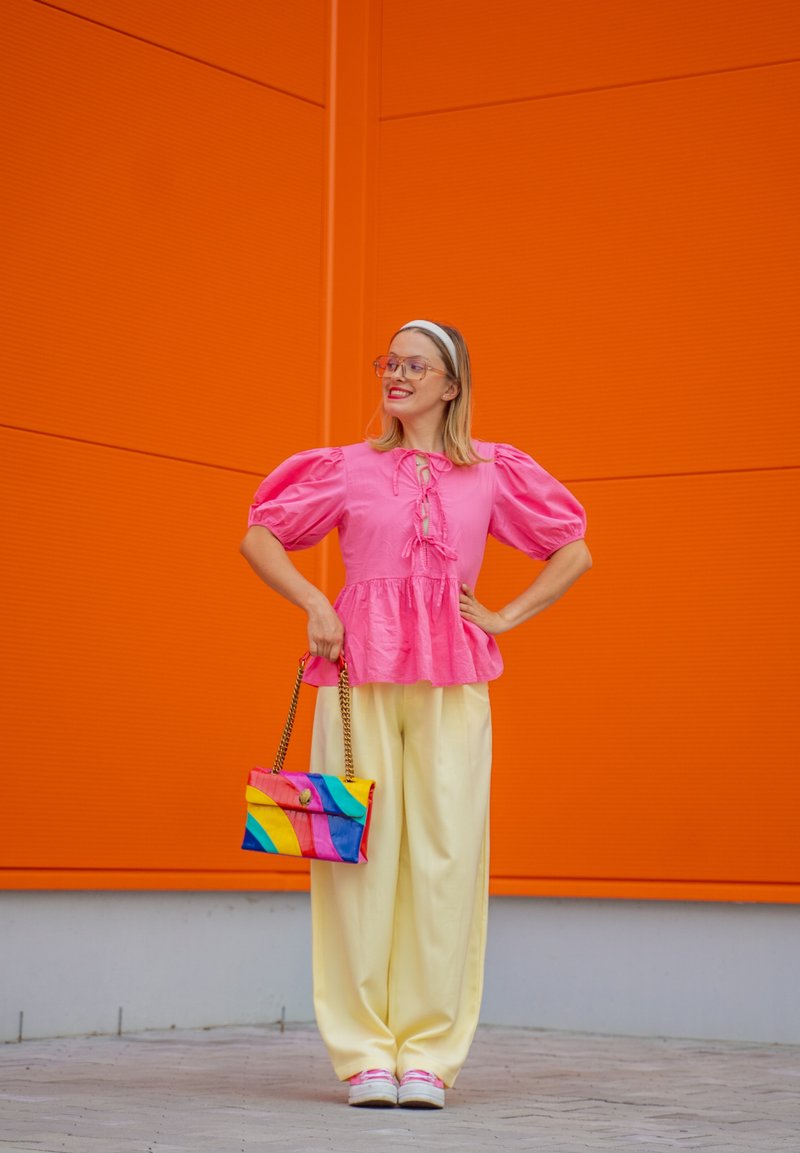 Blusa rosa com mangas bufantes e detalhe de laço, calças largas amarelas e uma mala listrada multicolorida. Fundo laranja brilhante.