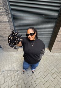 Femeie îmbrăcată cu mănuși și mâneci cu model de lună crescentă negre, tricou negru, blugi, pantofi negri și ochelari de soare mari, întinzându-se către cameră.