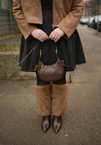 Femme portant une veste marron et des bottes en fourrure tenant un sac à main en cuir marron foncé avec des ferrures dorées sur un trottoir en ville.