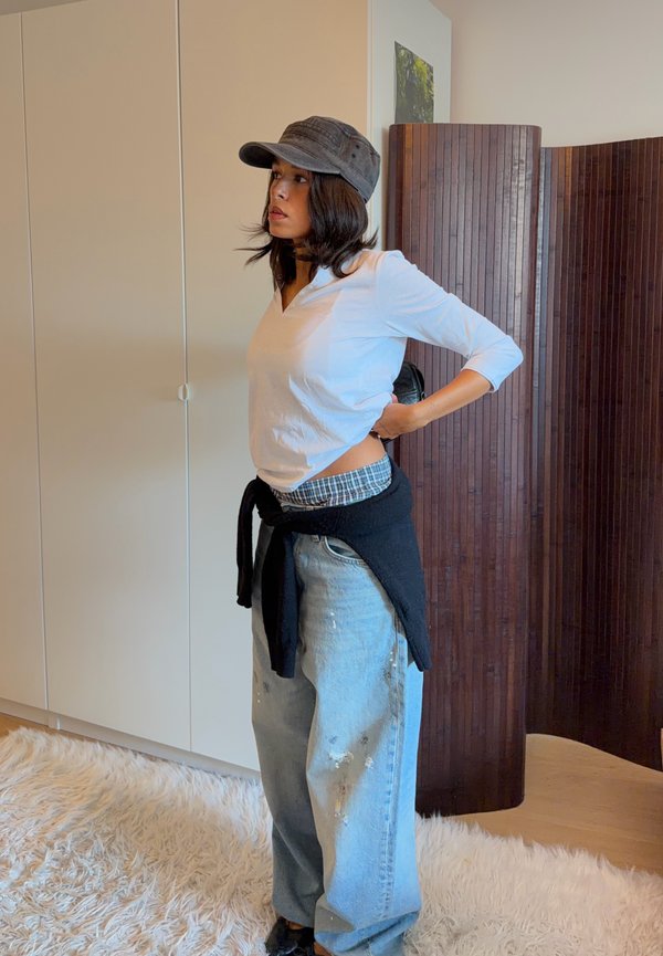 Mujer con gorra gris, camisa blanca de manga larga, vaqueros anchos y suéter negro atado a la cintura, de pie en el interior sobre una alfombra esponjosa.