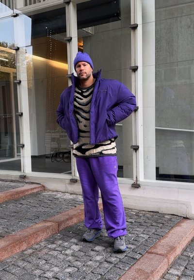 Hombre con gorro morado, chaqueta acolchada y pantalones de pie sobre un pavimento de adoquines frente a un edificio de vidrio y concreto.