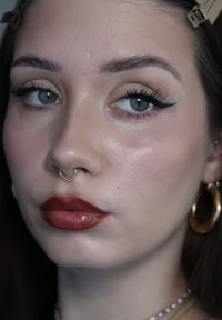 Gros plan sur une femme aux yeux bleus, avec un eye-liner ailé, un rouge à lèvres rouge foncé, un anneau de septum, une boucle d'oreille en or en forme d'anneau, et les cheveux attachés en arrière.