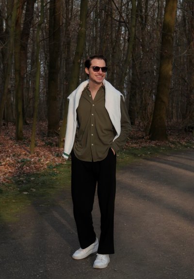 Persona vistiendo una camisa de botones verde oliva, pantalones amplios negros, zapatillas blancas y un suéter blanco caído sobre los hombros, en un bosque.