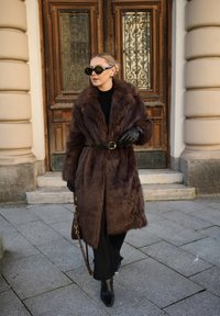 Manteau en fourrure marron avec un large col, ceinture noire cintrée, gants noirs et lunettes de soleil. Sol lisse et porte ornée en arrière-plan.