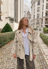 Trench-coat beige à double boutonnage porté sur une chemise blanche à boutons, accompagné d'un pantalon foncé ; cadre extérieur avec chemin pavé et végétation.