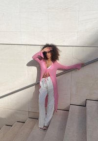 Lång rosa stickad omslag med klockärmar, knuten i midjan, i kombination med vita vida byxor och remsandaler i vitt på en trappa.