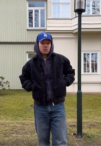 Jeune homme portant une casquette bleue de Los Angeles, une veste noire et un jean, debout à l'extérieur près d'un bâtiment et d'un lampadaire, les mains dans les poches.