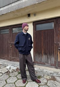 Jūras spēku bombers ar izšūtu dizainu, valkāts pār kreklu ar apkakli, kopā ar vaļīgām brūnām biksēm un vīnogu krāsas apaviem. Svītrainā cepure.