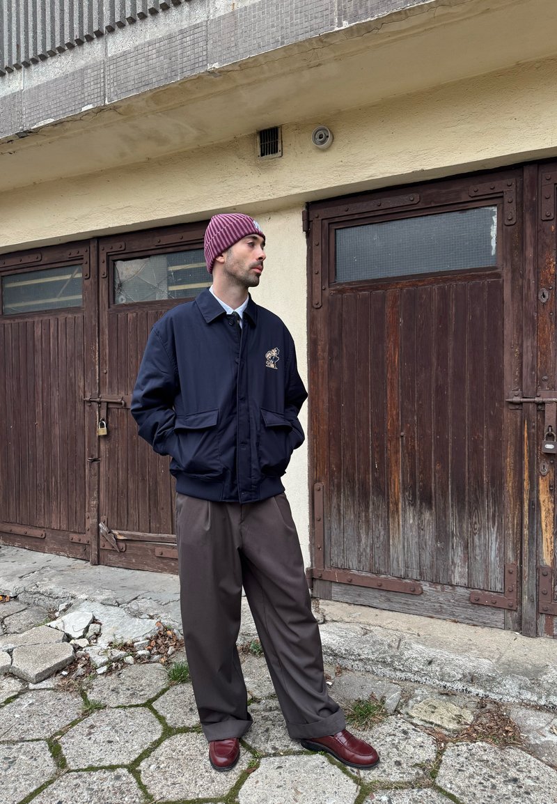 Veste aviateur bleu marine avec un motif brodé, portée sur une chemise à col, associée à un pantalon marron ample et des chaussures bordeaux. Bonnet rayé.