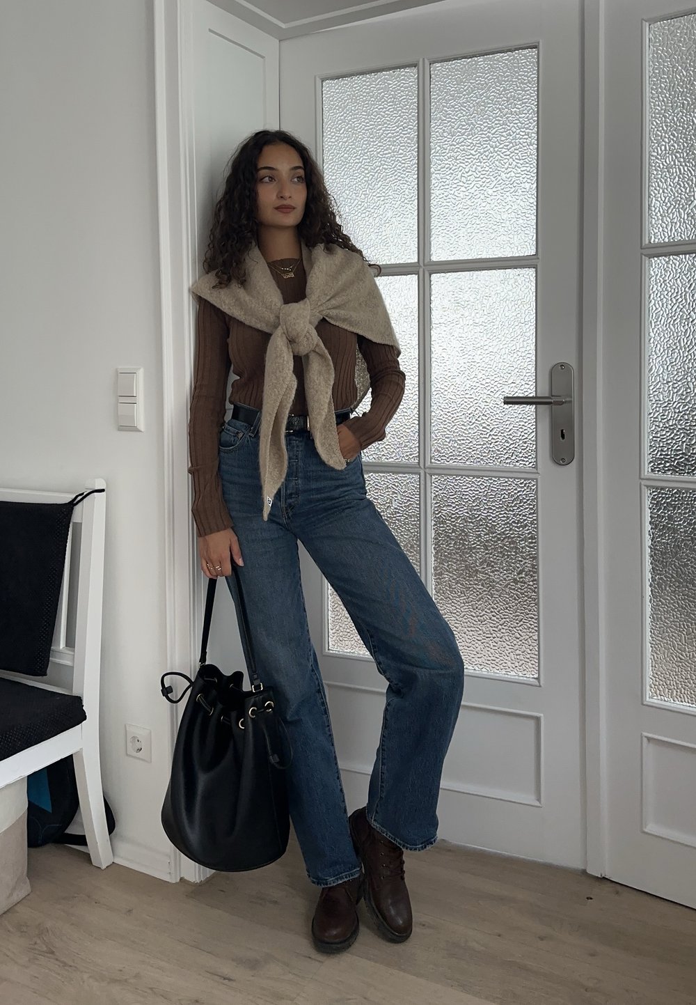 Brun ribstrikket langærmet top, beige striktrøje draperet over skuldrene, lyseblå jeans, brune ankelstøvler og en sort bucket-taske.