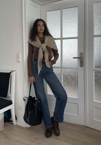 Top marrón de manga larga con textura acanalada, suéter beige de punto sobre los hombros, vaqueros azul claro, botines marrones y bolso tipo bucket negro.