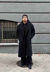 Jeune homme aux cheveux courts et foncés se tenant dans la rue, portant un long manteau noir, un pantalon noir et des chaussures noires, devant un mur gris.