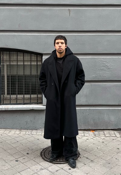 Joven con el pelo corto y oscuro de pie en la calle, vistiendo un abrigo negro largo, pantalones negros y zapatos negros, frente a una pared gris.