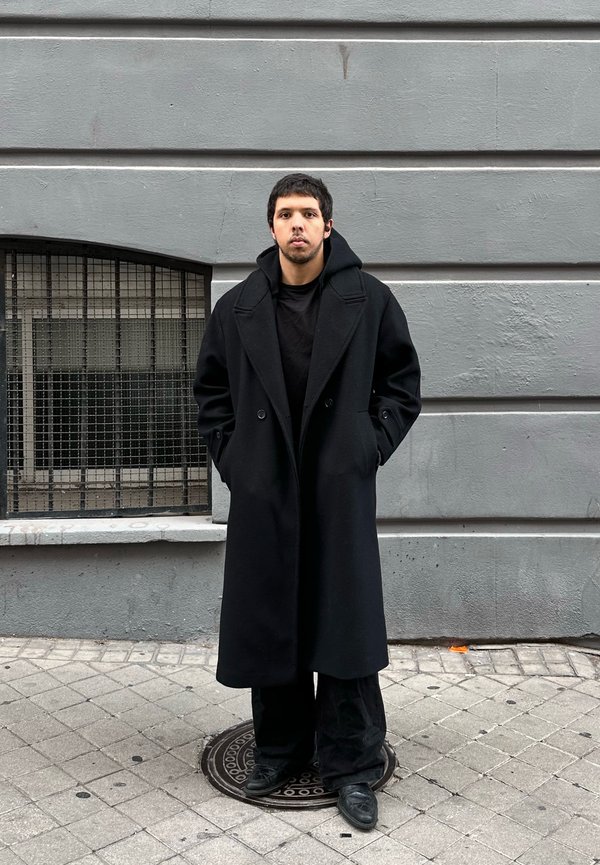 Mladić s kratkom tamnom kosom stoji na ulici noseći dugu crnu ogrtač, crne hlače i crne cipele na pozadini sive zidne površine.