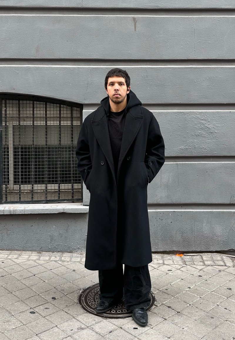 Jeune homme aux cheveux courts et foncés se tenant dans la rue, portant un long manteau noir, un pantalon noir et des chaussures noires, devant un mur gris.