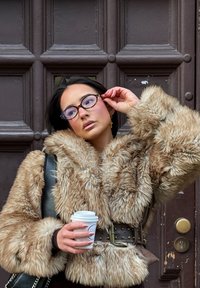 Brun korthåret pelsjakke med belte i midjen, grå stropp og naglede detaljer. Holder en kaffekopp, iført runde briller foran en mørk tremur.