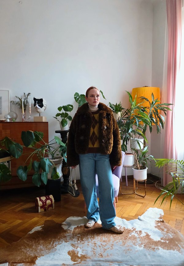 Femeie într-un palton maro de blană și blugi albaștri stă într-o cameră plină de plante, cu podea de lemn, covor de piele de vacă și perdele roz pal.