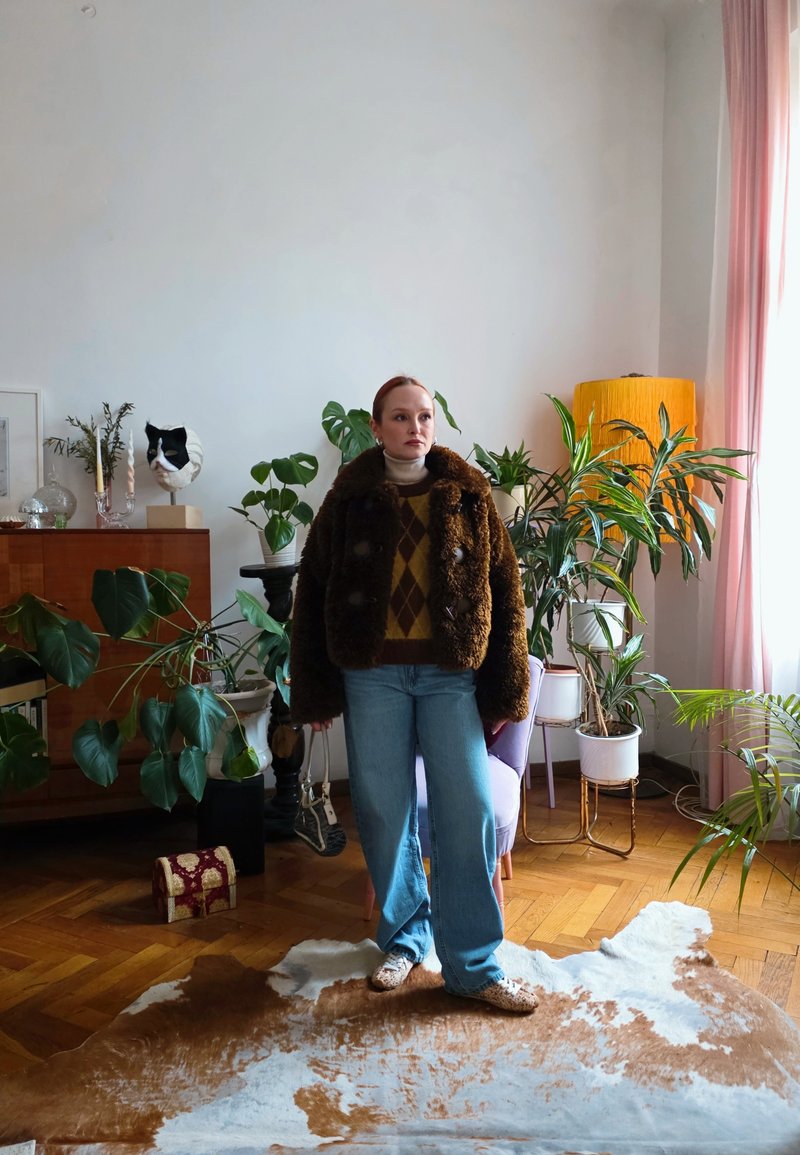 Femme en manteau de fourrure marron et jean bleu se tenant dans une pièce remplie de plantes avec un sol en bois, un tapis en peau de vache et des rideaux roses doux.