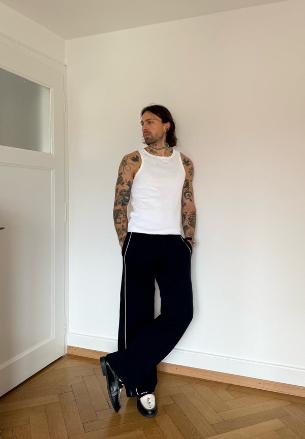 Mann med tatoverte armer iført hvit ermeløs skjorte, svarte bukser og svarte og hvite sko, som står foran en hvit vegg innendørs.