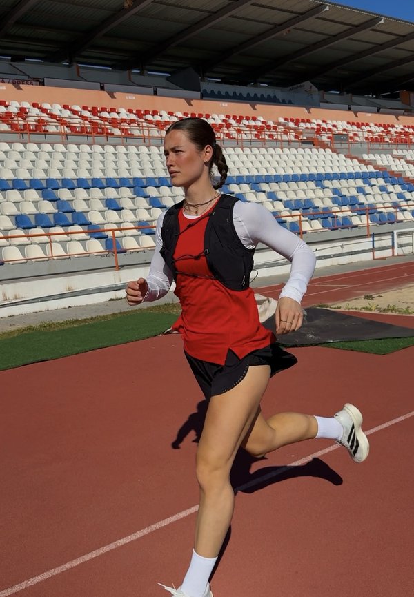 Női sportoló fut szabadtéri pályán, piros mellényben, fekete rövidnadrágban, fehér hosszú ujjú pólóban és futócipőben, üres stadion lelátói előtt.