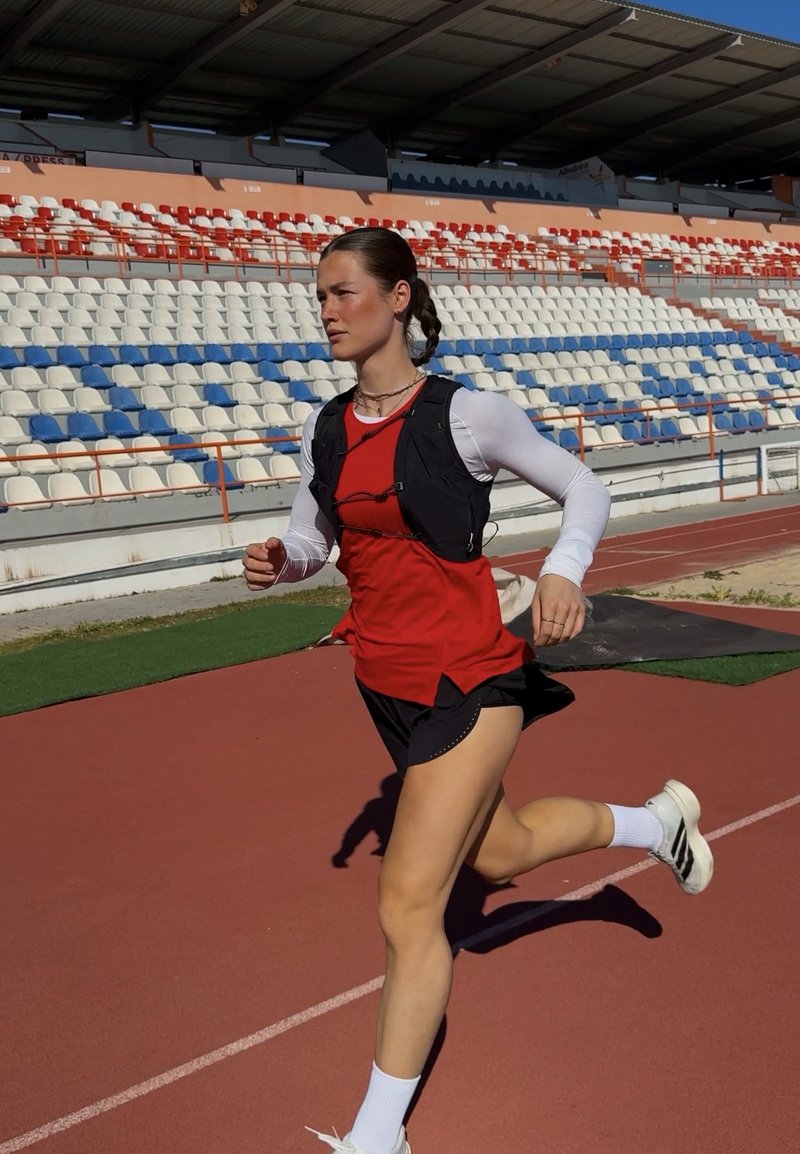 Atletă alergând pe pistă în aer liber, purtând vestă roșie, pantaloni scurți negri, bluză albă cu mâneci lungi și pantofi de alergare, în fața tribunei goale a stadionului.