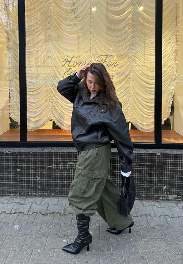 Młoda kobieta o długich włosach w czarnej skórzanej kurtce, oliwkowych spodniach typu cargo i czarnych botkach na obcasie idzie obok witryny sklepowej z zasłonami i napisem "Home For".
