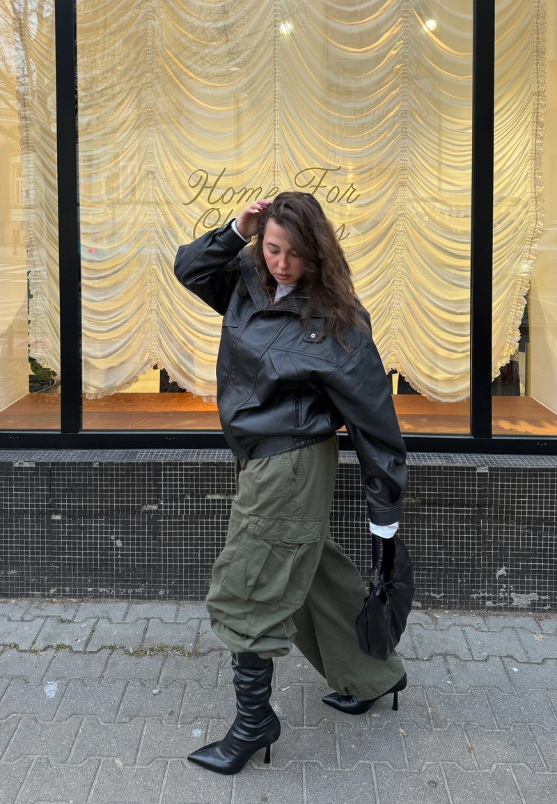 Junge Frau mit langen Haaren in schwarzer Lederjacke, olivgrünen Cargohosen und schwarzen Stiefeln mit Absatz, die an einem Schaufenster mit Vorhängen und der Aufschrift "Home For" vorbeigeht.
