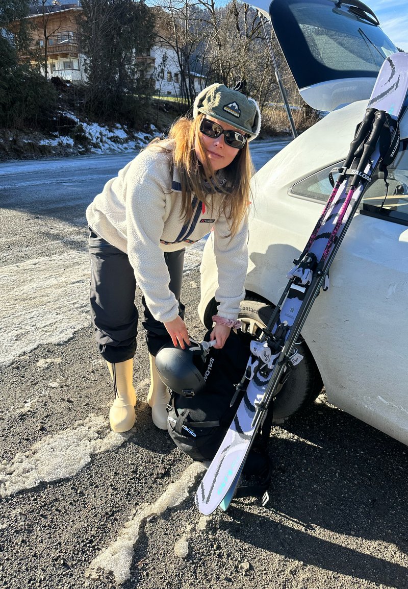 Scène d'équipement de ski : sac à dos noir posé au sol, skis blancs avec des touches de rose et de bleu appuyés contre une voiture, et une femme portant une veste en polaire.