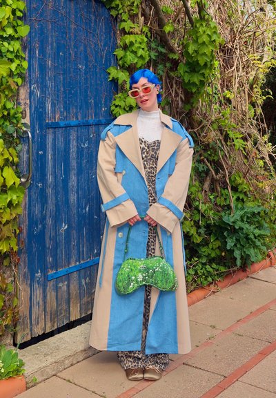 Abrigo largo beige y azul sobre un conjunto de estampado de leopardo, acompañado de un bolso verde con lentejuelas. Enmarcado por una puerta azul de madera y vegetación.
