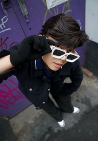 Bílé sluneční brýle s geometrickými obroučkami, nošené s tmavou bundou a rukavicemi, pózující před zdí pokrytou fialovým graffiti. Viditelné jsou pruhované kalhoty.