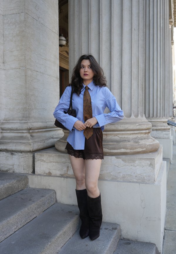 Joven mujer con camisa azul de rayas, corbata con estampado, pantalones cortos negros y botas, de pie en escalones de piedra junto a grandes columnas clásicas.