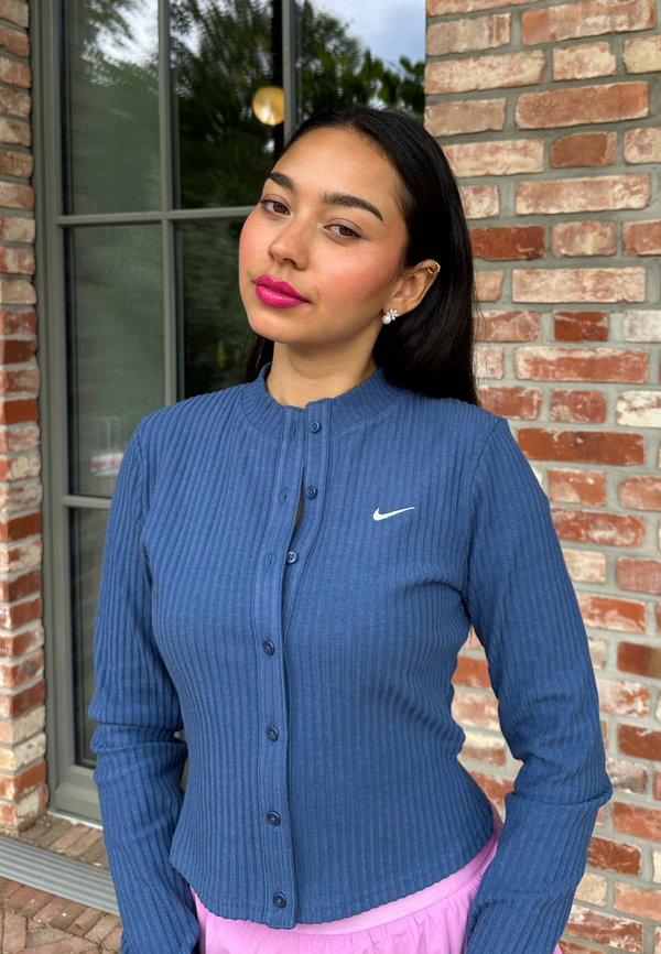 Young woman with long dark hair wearing a blue Nike buttoned cardigan and pink skirt standing in front of a brick wall and window.