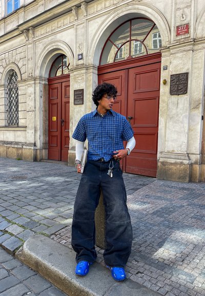 Camisa de cuadros azul de manga corta, camiseta blanca de mangas largas, vaqueros negros de gran tamaño, zapatillas azules, sosteniendo un portátil, con llaves y accesorios.
