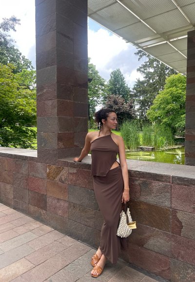 Vestido marrón con un corte asimétrico, textura suave y un detalle de corte. Los accesorios incluyen un bolso blanco texturizado y calzado marrón.