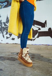 Botas castanhas forradas a pelo, com um exterior texturizado, apresentando um design de atacadores e uma sola grossa cor de tan, combinadas com jeans azuis e um casaco amarelo.