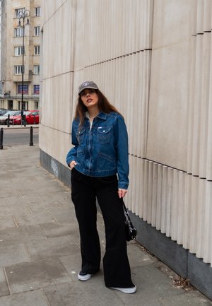 Chaqueta de denim en azul con bolsillos delanteros, combinada con pantalones de pierna ancha negros. Los accesorios incluyen una gorra gris y un pequeño bolso negro.