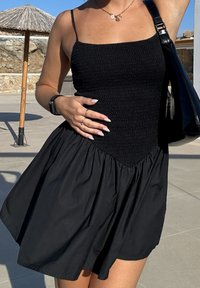 Črna poletna obleka s prilegajočim se zbranim zgornjim delom in razširjenim krilom. Tanke naramnice, gladka tekstura in priložnostni slog. Vidna črna torbica in pametna ura.