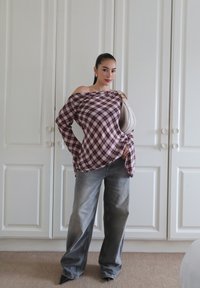 Crvena i bijela karirana off-shoulder majica, svijetlosivi široki traperice i crne špicaste potplate. Bež torba s jednim remenom. Bijela pozadina.