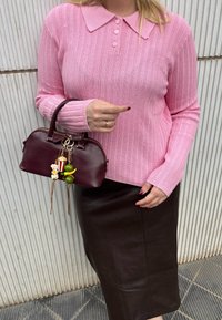 Różowy sweter o prążkowanej strukturze z kołnierzykiem, zestawiony z brązową skórzaną spódnicą midi. Trzymając małą bordową torebkę z dekoracyjnymi zawieszkami.
