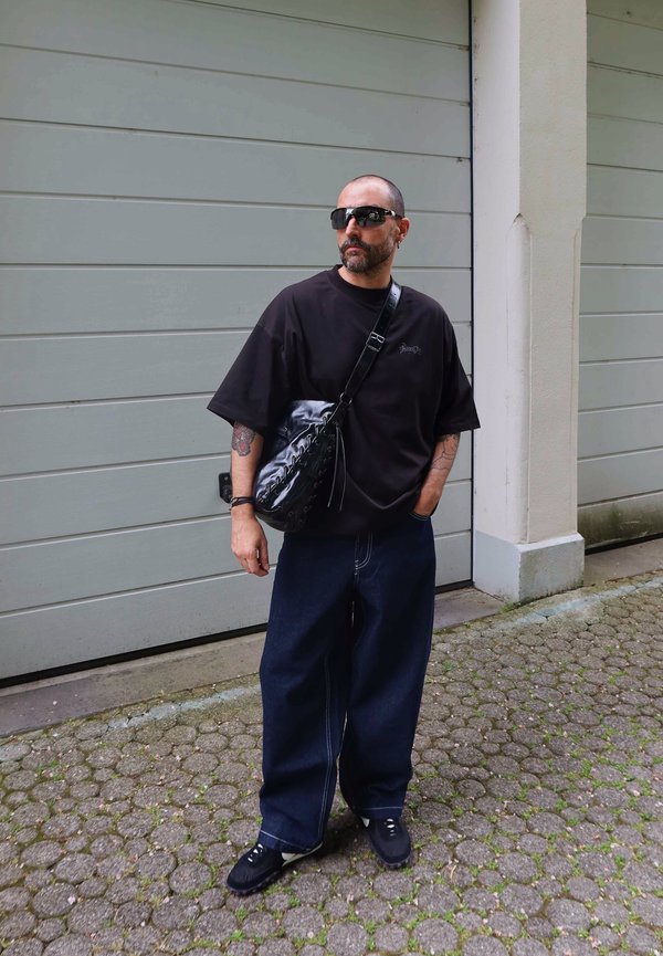 Mand med barberet hoved og skæg iført sort oversized t-shirt, brede mørke jeans, sorte sneakers, solbriller og bærer en sort crossbody taske.