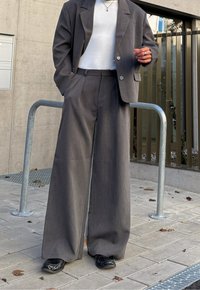 Personne portant un pantalon de costume gris à jambes larges et un blazer assorti sur un haut blanc, debout sur une surface pavée près d'une rampe métallique.