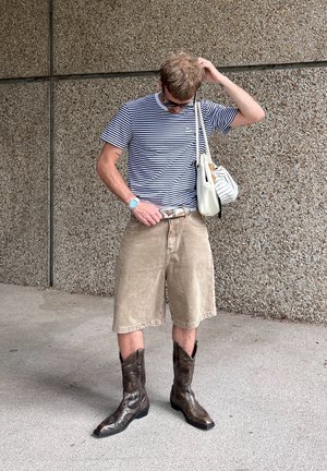 Camiseta de rayas con cuello redondo, pantalones cortos de mezclilla beige y botas de vaquero marrones. Los accesorios incluyen un bolso blanco y un reloj azul.