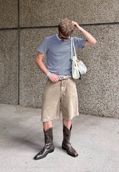 Camiseta de rayas con cuello redondo, pantalones cortos de mezclilla beige y botas de vaquero marrones. Los accesorios incluyen un bolso blanco y un reloj azul.